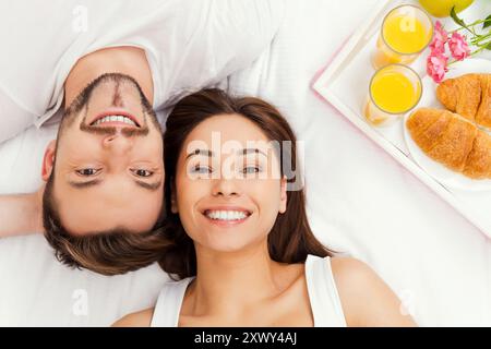 Joyeux matin. Vue de dessus de beau jeune couple aimant couché dans le lit ensemble et souriant tandis que plateau avec petit déjeuner couché près d'eux Banque D'Images