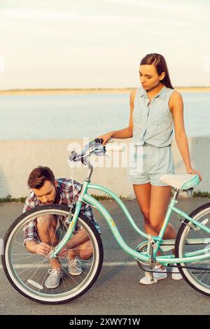 Je peux compter sur lui dans n'importe quelle situation. Jeunes femmes frustrées debout derrière son vélo alors que l'homme réparait une roue Banque D'Images