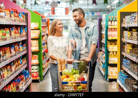 Profiter du shopping ensemble. Heureux jeune couple se liant l'un à l'autre et souriant en marchant tout en marchant dans un magasin d'alimentation avec panier Banque D'Images