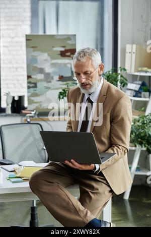 Un homme mûr avec une barbe est assis sur un bureau, regardant attentivement son ordinateur portable, tout en enseignant en ligne. Banque D'Images