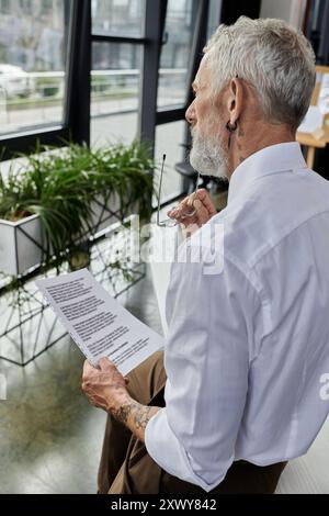Un professeur LGBTQ mature avec une barbe lit un document tout en enseignant en ligne. Banque D'Images