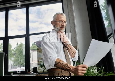 Un homme d'âge moyen avec une barbe et des lunettes lit un papier tout en se préparant pour un cours en ligne. Banque D'Images