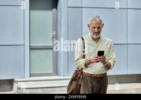 Un homme d'âge mûr avec une barbe vérifie son téléphone en marchant à l'extérieur d'un bâtiment moderne. Banque D'Images