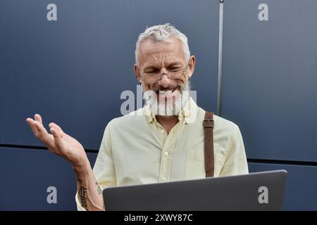 Un homme barbu dans une chemise jaune et des lunettes sourit en utilisant un ordinateur portable, la main gauche levée en l'air. Banque D'Images