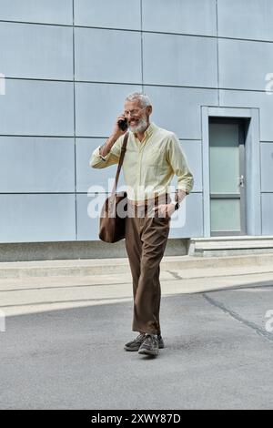 Un homme d'âge mûr avec une barbe sourit tout en parlant sur son téléphone à l'extérieur d'un bâtiment moderne. Banque D'Images