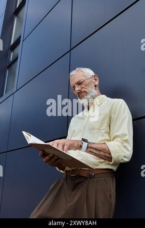 Un homme gay d'âge moyen avec une barbe lit un livre tout en se tenant devant un bâtiment moderne. Banque D'Images