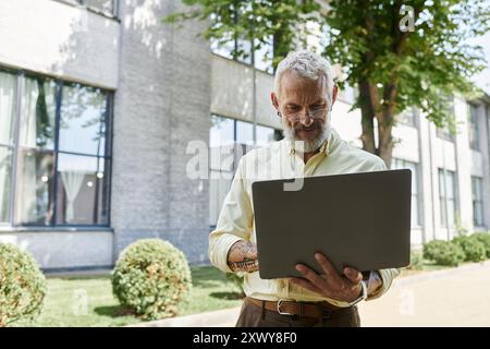 Un homme d'âge moyen avec une barbe et des lunettes utilise son ordinateur portable à l'extérieur d'un bâtiment moderne. Banque D'Images