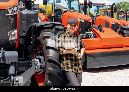 Représentant commercial féminin dans une concession de machines agricoles et de construction se tient à côté d'un tracteur tient une tablette numérique et souriante. Banque D'Images