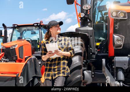 Femme représentante dans une concession pour la construction et les machines agricoles se tient à côté d'un tracteur tient tablette numérique et souriant. Banque D'Images