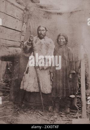 Peuples autochtones minoritaires du Nord, de Sibérie et de l'extrême-Orient de la Russie. Nivkh femme et homme debout près d'un mur de bois (groupe habitant la moitié nord de l'île Sakhaline). Un homme vêtu d'un long manteau de fourrure tient une lance dans ses mains. Une femme en vêtements simples sombres et de longues bottes. De l'expédition au Kamtchatka et aux îles commander par le célèbre naturaliste et médecin polonais. Benedykt Dybowski. Empire russe. années 1870 Banque D'Images