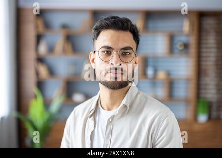 Portrait en gros plan d'un jeune indien portant des lunettes et une chemise de couleur claire assis sur le canapé à la maison, regardant sérieusement et avec confiance la caméra. Banque D'Images