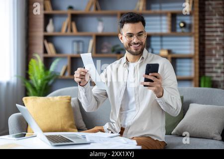 Jeune homme indien travaillant à la maison avec un ordinateur portable et des factures, tenant le reçu et regardant souriant l'écran du téléphone portable, vérifiant les documents. Banque D'Images