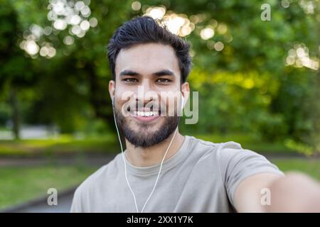 Jeune homme souriant et prenant selfie avec téléphone dans le parc extérieur. Profiter de la nature et écouter de la musique avec des écouteurs. Vêtements décontractés et expressions joyeuses. Banque D'Images