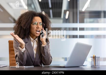Jeune femme d'affaires afro-américaine en colère assise dans le bureau au bureau et parlant émotionnellement sur le téléphone portable. Banque D'Images