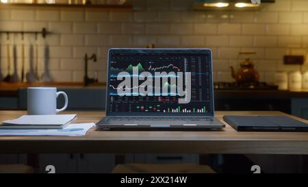 Un ordinateur portable affichant des graphiques financiers et des données boursières sur une table en bois dans une cuisine. Trader à domicile ou analyste, concept financier Banque D'Images