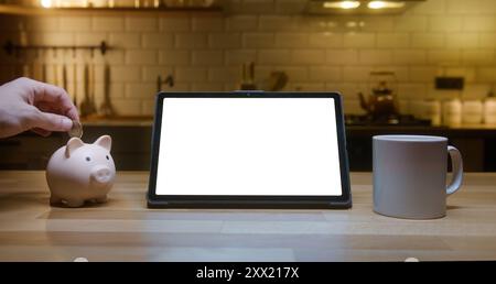 Ordinateur tablette avec écran blanc maquette chromakey debout sur la table de cuisine la nuit. Une personne inconnue met de l'argent dans une tirelire. Sa Banque D'Images