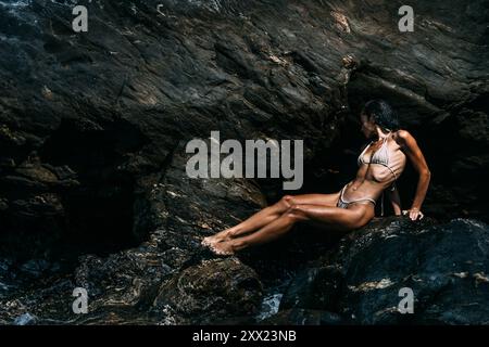 Vue de côté d'une femme athlétique dans un bikini assis sur les rochers côtiers, Espagne Banque D'Images