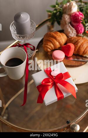 Gros plan d'un bouquet de fleurs et d'un coffret cadeau sur une table avec une tasse de café, des croissants frais et des macarons pour un petit déjeuner Saint-Valentin Banque D'Images