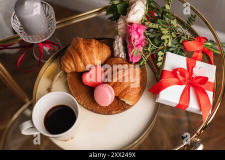 Vue aérienne d'un bouquet de fleurs et coffret cadeau sur une table avec une tasse de café, des croissants frais et des macarons pour un petit déjeuner Saint-Valentin Banque D'Images