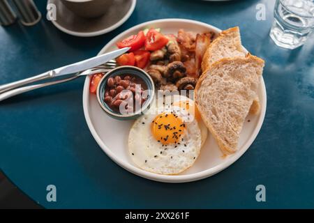 Petit-déjeuner anglais traditionnel avec haricots, bacon, œufs frits, champignons, tomates, concombre et pain avec un cappuccino et un verre d'eau Banque D'Images