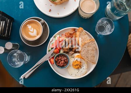 Petit-déjeuner anglais traditionnel avec haricots, bacon, œufs frits, champignons, tomates, concombre et pain avec un cappuccino et un verre d'eau Banque D'Images