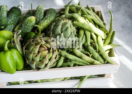 Gros plan d'artichauts frais, de haricots secs, de concombres et de poivrons verts dans une caisse en bois Banque D'Images