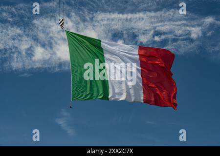 Un grand drapeau italien ondulant dans le ciel soutenu par un crochet de grue. Composition horizontale et espace de copie. Banque D'Images