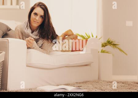 La jeune femme est allongée sur le ventre sur un canapé blanc avec les pieds en l'air, elle porte un pull beige et une écharpe Banque D'Images