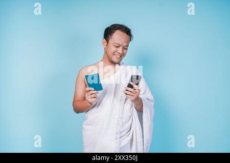 Joyeux jeune homme musulman asiatique portant un ihram tenant un passeport tout en regardant son smartphone sur fond bleu isolé. Hajj concept. Banque D'Images