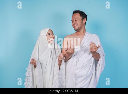 Jeune couple musulman asiatique souriant portant ihram et robe de prière avec geste de la main de prière sur isolé par fond bleu. Hajj concept. Banque D'Images