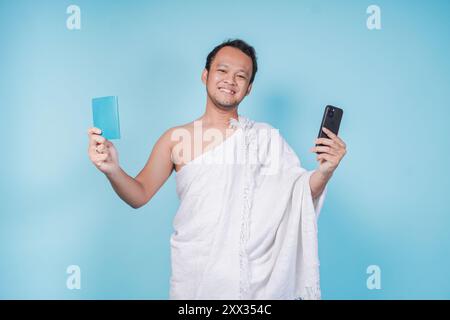 Joyeux homme musulman asiatique portant ihram tenant smartphone et passeport avec un sentiment excité, fond bleu isolé. Hajj concept. Banque D'Images