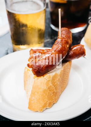 Saucisse de chorizo sur une tranche de pain sur une assiette blanche, accompagnée d'un verre de bière dans un cadre décontracté. Banque D'Images