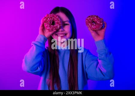 Photo d'adorable petite fille porter un sweat à capuche mangeant des beignets glacés espace vide isolé fond de couleur bleu magenta Banque D'Images