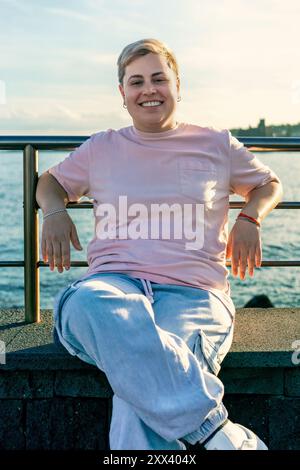 Femme de grande taille joyeuse avec les cheveux courts et piercing nez est assis sur la rampe de front de mer, profitant du coucher du soleil. Positivité corporelle, acceptation de soi et diversification Banque D'Images