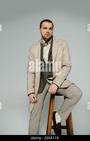 Un homme élégant portant un manteau beige et des chaussures marron est assis sur un tabouret sur un fond gris. Banque D'Images