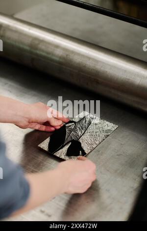 Angle élevé de jeunes mains artisanales mettant des œuvres d'art sur le plateau en acier de la machine d'impression par rouleau tout en travaillant dans la typographie Banque D'Images