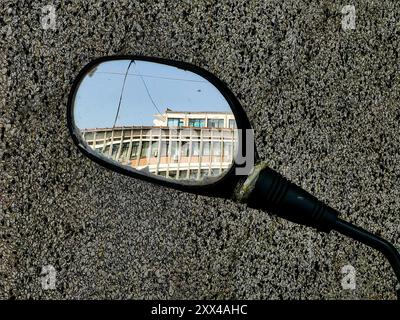 Réflexions du passé : dans le miroir de vue latérale endommagé d'un scooter reflétant un ancien bâtiment de l'hôpital contre un mur de granit - jour ensoleillé avec Blue Sky Banque D'Images