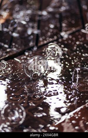 Gros plan des verres à vin cassés et du vin renversé, des parties du verre cassé sur le sol avec des éclaboussures d'eau sur la terrasse en bois Banque D'Images