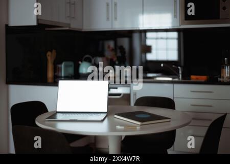 Une photo discrète d'un ordinateur portable avec un écran blanc vierge sur une table ronde dans une cuisine moderne, accompagnée d'un cahier, d'un stylo et d'un éléme de décor minimaliste Banque D'Images