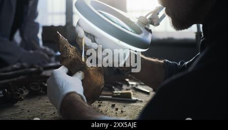 Paléoanthropologue ou archéologue mature nettoie les restes fossiles avec une brosse, étudie les os de l'homme ou de l'animal antique sous la loupe d'une lampe dans un laboratoire archéologique. Scientifique travaillant en arrière-plan. Banque D'Images