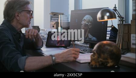 Scientifique mature ou chercheur étudie le crâne fossile, utilise une tablette et un ordinateur avec visualisation 3D de la tête humaine éteinte ancienne, prend des notes dans un cahier. Archéologue travaille dans un laboratoire archéologique. Banque D'Images