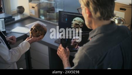 Deux archéologues étudient le crâne fossile, créent un modèle 3D de la tête humaine préhistorique éteinte sur tablette et ordinateur en utilisant la technologie de visualisation. Les scientifiques discutent, travaillent dans un laboratoire archéologique. Banque D'Images