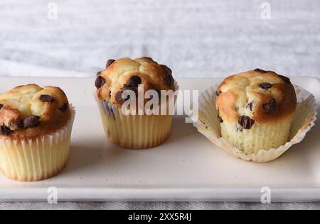 Gros plan de trois mini muffins aux pépites de chocolat sur une assiette de service blanche et un torchon en lin. Banque D'Images