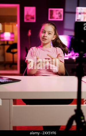 L'enfant joyeux utilise le téléphone portable sur le stabilisateur de trépied pour tirer le segment d'introduction pour le vlog. Jeune créateur de contenu dans le studio rose néon accueille la communauté de fans dévouée, filmant avec un téléphone portable Banque D'Images