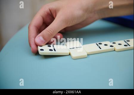 Gros plan d'une main plaçant habilement une tuile de domino sur une table bleue, mettant en évidence la stratégie et la concentration dans le jeu classique. Banque D'Images