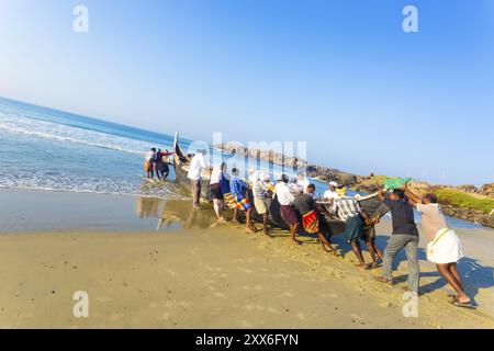 Effort communautaire d'aide des villageois poussant le bateau de pêche ensemble de la plage de sable dans l'eau un matin ensoleillé à Kovalam, Kerala, Inde, Asie Banque D'Images