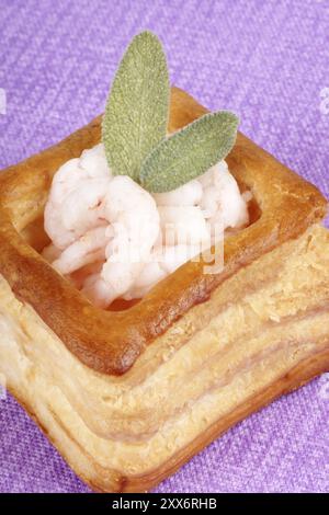 Close-up of a squared vol-au-vent farci de petites crevettes cuites décorées avec des feuilles de sauge sur fond violet Banque D'Images