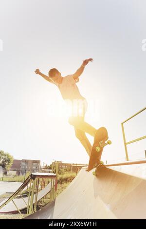 Adolescent patineur raccroché au-dessus d'une rampe sur une planche à roulettes dans un skate Park au coucher du soleil. Grand angle. Image chaude ensoleillée Banque D'Images