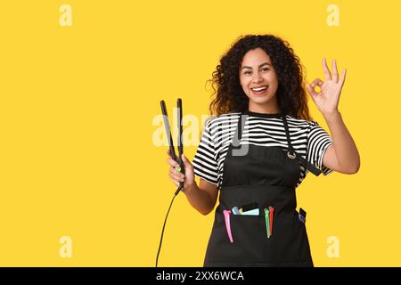 Beau jeune heureux coiffeur afro-américain avec lisseur de cheveux montrant un geste OK sur fond jaune Banque D'Images