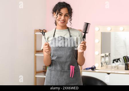 Belle jeune coiffeuse afro-américaine heureuse avec ciseaux, brosse et pinces à cheveux dans le salon de beauté Banque D'Images